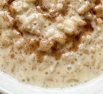 Bol de arroz con leche cremoso con canela en polvo y una rama de canela, servido en cerámica blanca sobre mesa de madera